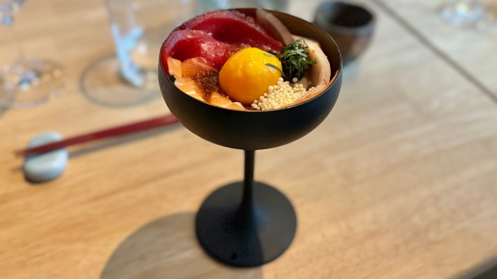 A beautifully arranged sashimi cocktail in a black bowl, featuring assorted seafood, a bright egg yolk, and garnishes, placed on a wooden table with chopsticks and glasses in the background.