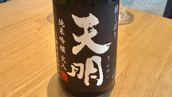 A close-up of a bottle of sake labeled 'Tenmei' on a wooden table, with a soft background and a wine glass partially visible.