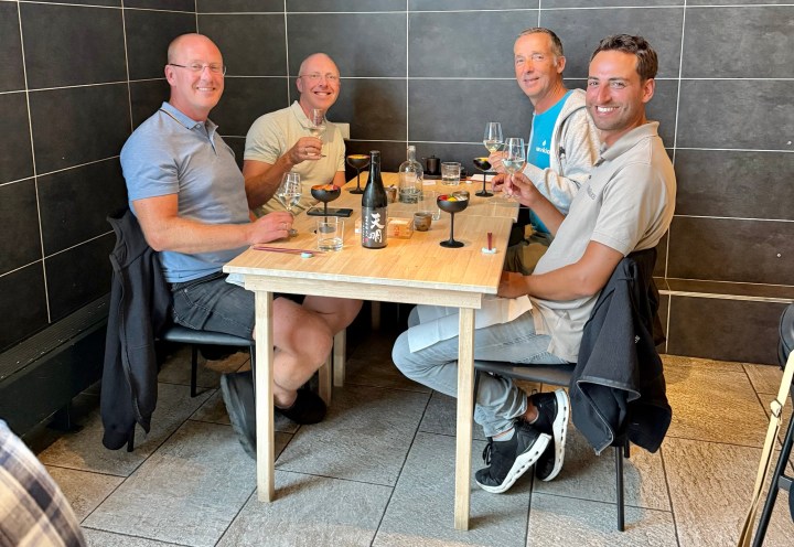 Four men sitting at a table in a restaurant, raising glasses in a toast. The table is set with various drinks and small dishes, and the interior features dark tiled walls and a minimalist design.