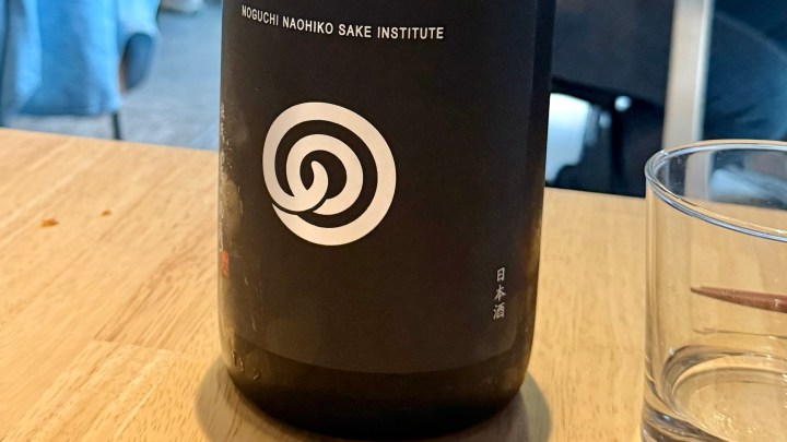 A bottle of sake from the Noguchi Naohiko Sake Institute, featuring a spiral logo, placed on a wooden table beside a glass.