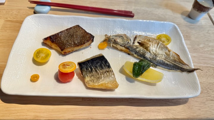 A beautifully arranged plate of grilled fish, featuring different types including mackerel and a whole fish, garnished with colorful vegetables like lemon, cherry tomatoes, and shiso leaves, served on a textured white plate with red chopsticks in the background.