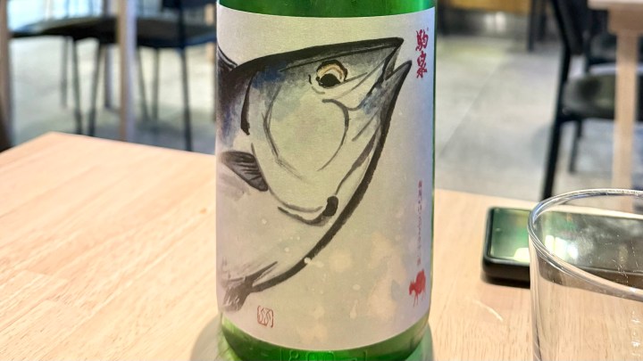 A close-up of a sake bottle featuring an illustration of a fish, placed on a wooden table in a restaurant setting.