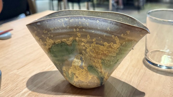 A unique metallic sake cup with an artistic design, placed on a wooden table in a restaurant setting.