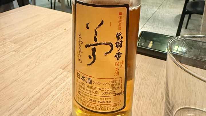 A close-up of a bottle of sake with a distinctive label, placed on a wooden table next to a glass. The label features intricate Japanese characters and details about the sake.