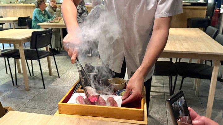 A chef finishing wagyu nigiri at Hokkai Kitchen, with smoke rising from the dish, surrounded by diners and empty tables.