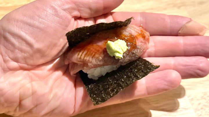 A hand holding a piece of sushi, featuring a slice of wagyu beef topped with wasabi and served on rice, wrapped in a sheet of nori.