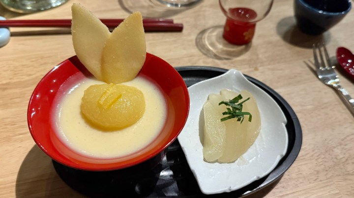 A dessert featuring custard topped with nashi pear marinated in yuzu juice, garnished with shiso leaves, served in a red bowl, alongside a small dish of additional marinated pear.