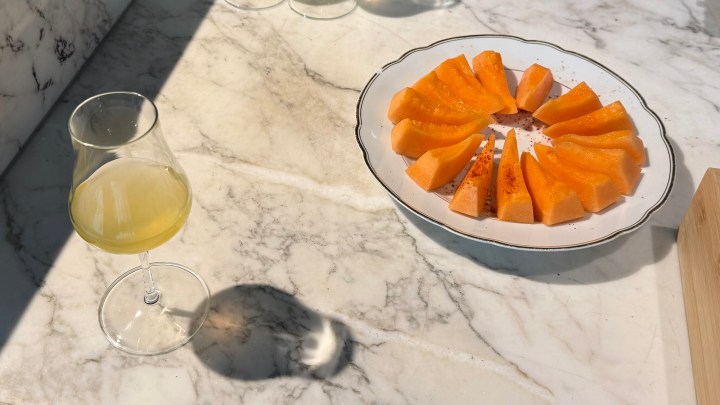 A glass of kombucha next to a plate of cantaloupe slices sprinkled with Moroccan spices, arranged on a marble table.