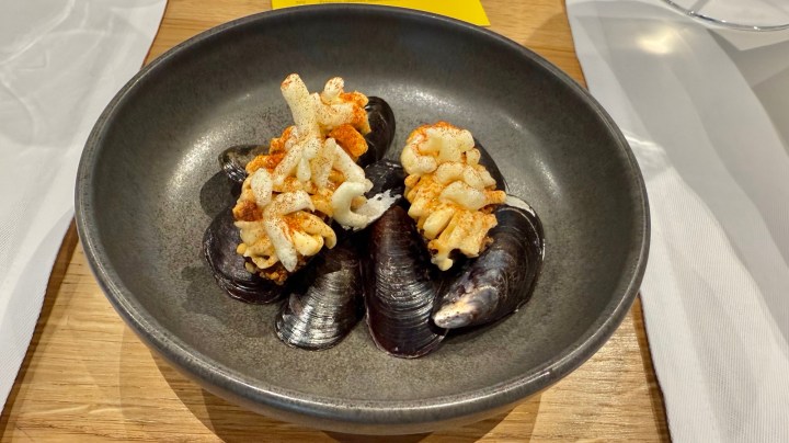 A plate featuring mussel shells filled with a creamy mussel and herb mixture, topped with a crunchy garnish, served on a wooden table.