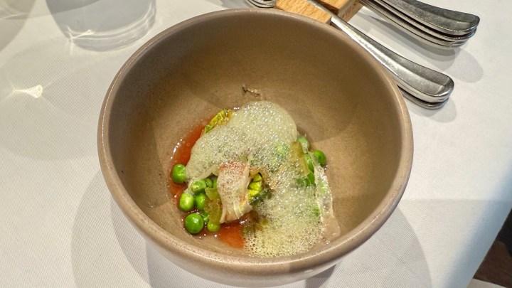 A bowl of sweet, crunchy peas with basil, tomato, and a light foam, presented in a stylish restaurant setting.