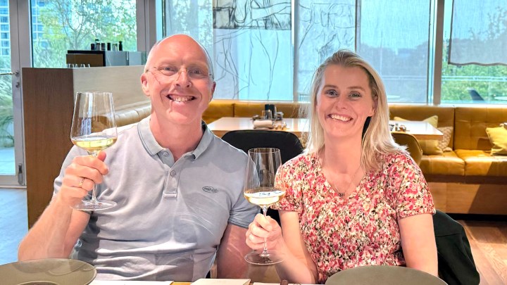 A man and a woman holding glasses of white wine, smiling at the camera while seated at a restaurant table with a cozy ambiance in the background.