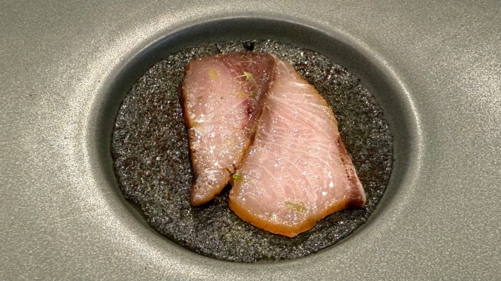 Close-up of cured hamachi belly served on a dark plate, showcasing two slices of fish with a glossy appearance.