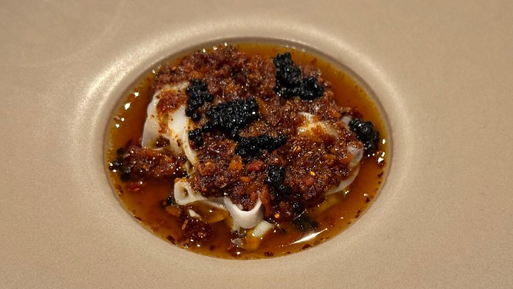 A gourmet dish featuring thinly sliced cuttlefish topped with a blend of crunchy texture and dark seasoning in a savory broth, elegantly presented in a round bowl.