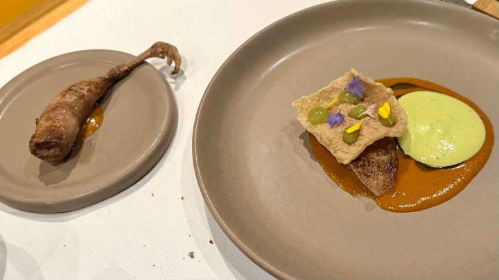 A plated dish featuring a quail leg alongside a creamy sauce and a textured component, garnished with colorful edible flowers.