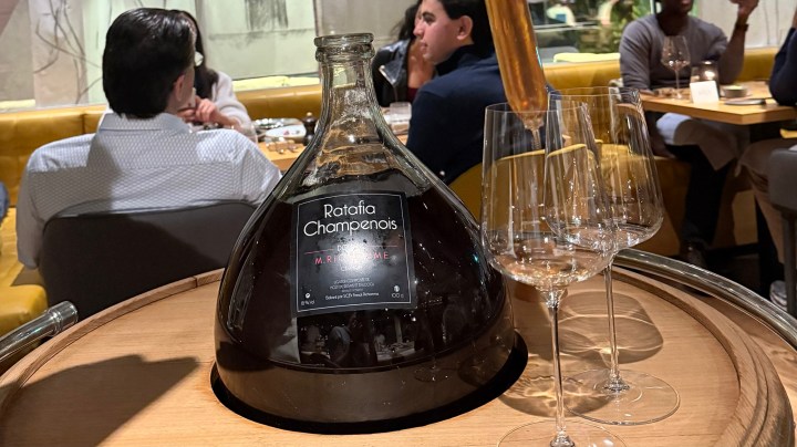 A large bottle of Ratafia Champenois displayed on a wooden tray, accompanied by three empty glasses, with restaurant patrons visible in the background.