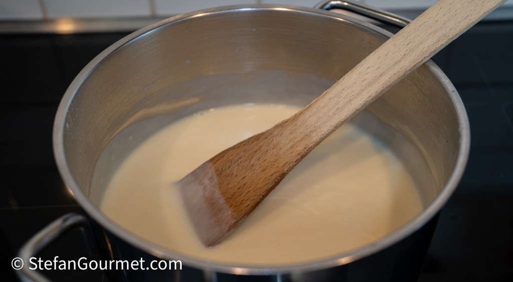 A saucepan containing a creamy white sauce with a wooden spoon resting inside.
