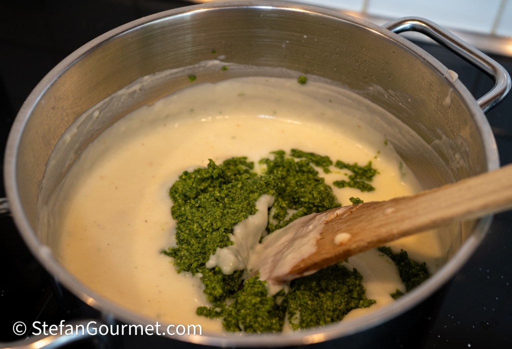 A pot on a stove containing a creamy white sauce with fresh green pesto being mixed in with a wooden spoon.
