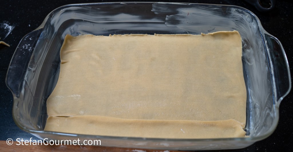 A greased baking dish with fresh pasta dough laid out at the bottom, ready for lasagna preparation.