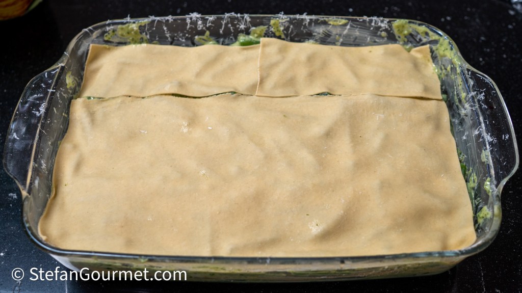 A glass baking dish filled with layers of fresh pasta sheets and a green pesto mixture, topped with more pasta, ready for baking.