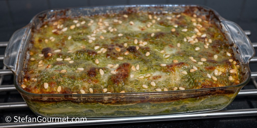 Baked lasagna alla genovese topped with melted cheese and pine nuts in a glass dish.