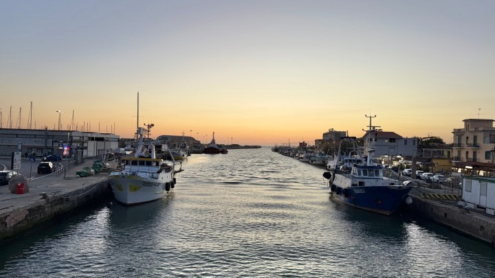 View of a fishing harbor at sunset, with boats docked along the shore and the water reflecting the warm colors of the sky.