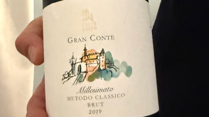 Close-up of a hand holding a bottle of Gran Conte 2019 Brut wine, featuring an illustrated label with a castle and the text 'Gran Conte Millesimato Metodo Classico Brut'.