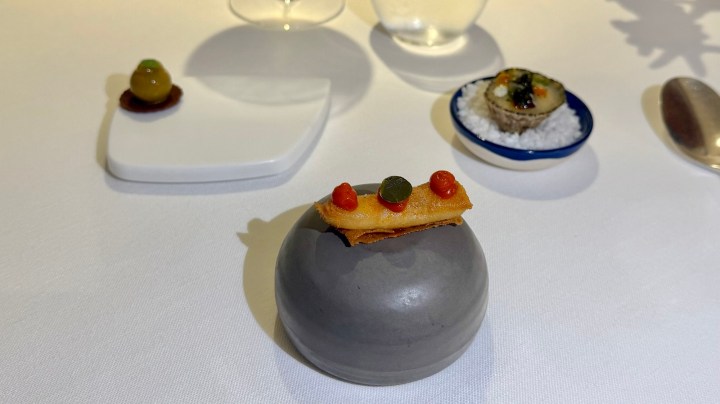 A gourmet amuse-bouche presented on a round gray stone, featuring a delicate crispy layer topped with colorful small dots, alongside minimalist white plates and a blue dish with seafood on a bed of salt.