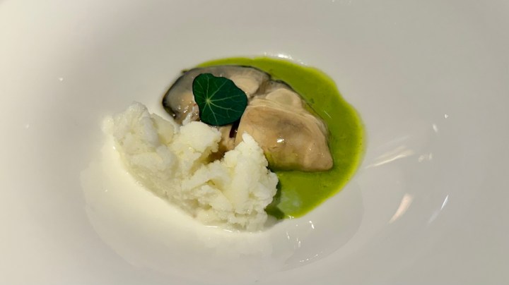 A plated dish featuring a French oyster served with a white chocolate granita and a vibrant green sauce, garnished with a small leaf.