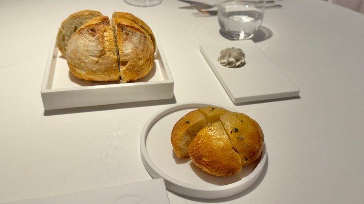 An elegantly set dining table featuring a sliced round sourdough loaf and a smaller bun called focaccina, accompanied by a small portion of whipped butter on a white plate.
