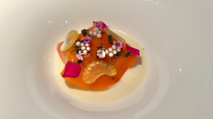 A beautifully presented dish featuring a bright orange ravioli adorned with edible flowers and decorative garnishes, served in a creamy sauce.