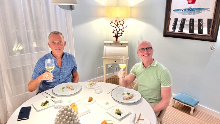 Two men smiling at a dining table in a restaurant, each holding a glass of wine, with a light and airy decor in the background.