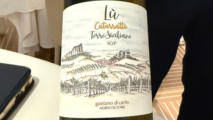 Close-up of a wine bottle labeled 'Lù Catarratto Terre Siciliane IGP' with a scenic illustration of vineyards and hills.