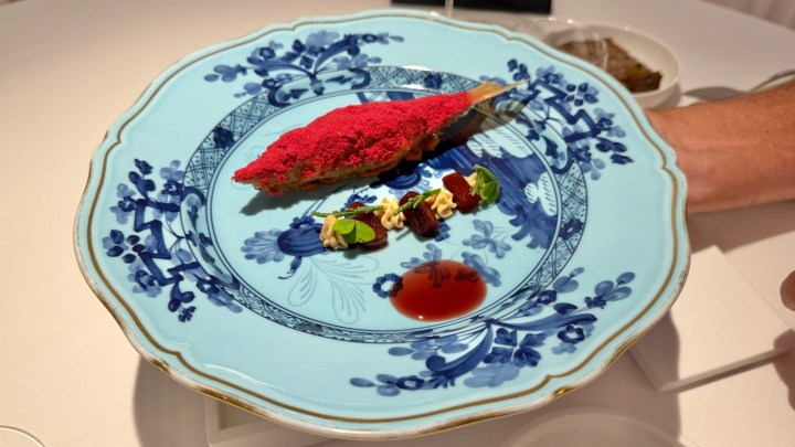 A beautifully presented dish featuring a fish topped with a vibrant red powder, accompanied by colorful garnishes and a sauce, served on a blue decorative plate.