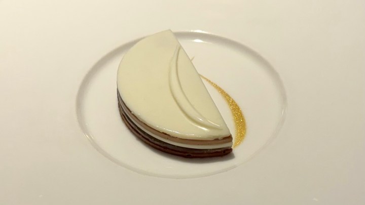 A beautifully plated dessert featuring a layered cake with white and dark chocolate, shaped in a crescent, resting on a white plate with a golden decorative element.