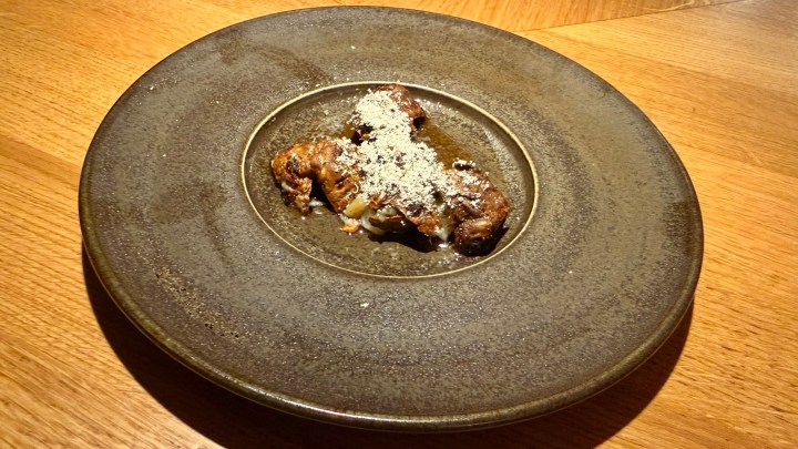 A beautifully plated dish featuring a piece of food topped with grated cheese or a similar ingredient, served on a rustic, dark ceramic plate with a textured finish, against a warm wooden background.