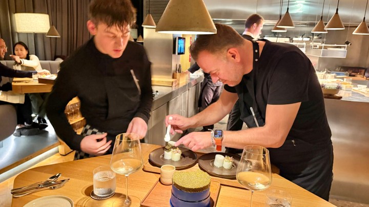 Two chefs in a restaurant kitchen, one finishing a dish on a plate while the other assists, with glasses of wine and other plates on the table.