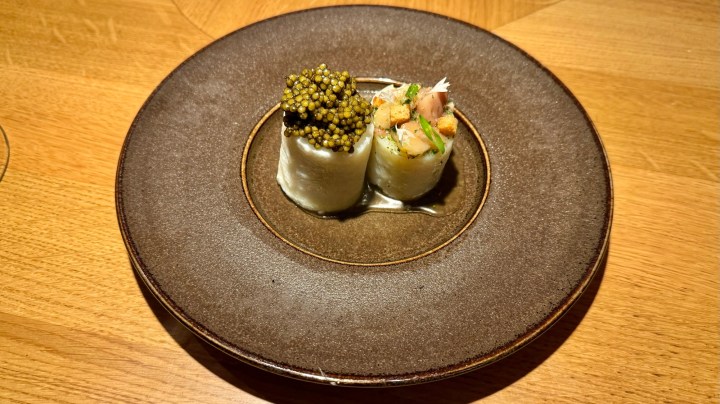 A gourmet dish featuring rolled fish garnished with green caviar, served on a dark plate.