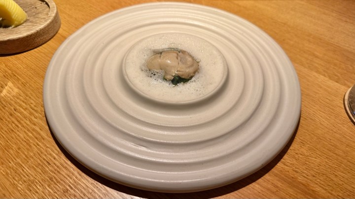 A beautifully presented dish featuring an oyster on a white, textured plate, surrounded by green sauce and topped with frothy foam.