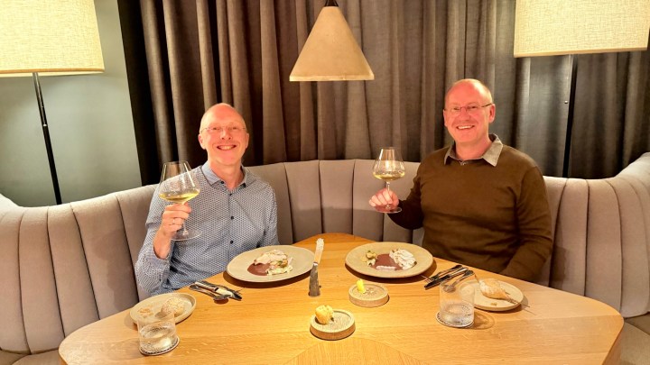 Two men seated at a restaurant table, each holding a glass of wine while smiling. Plates of food are in front of them on a wooden table.