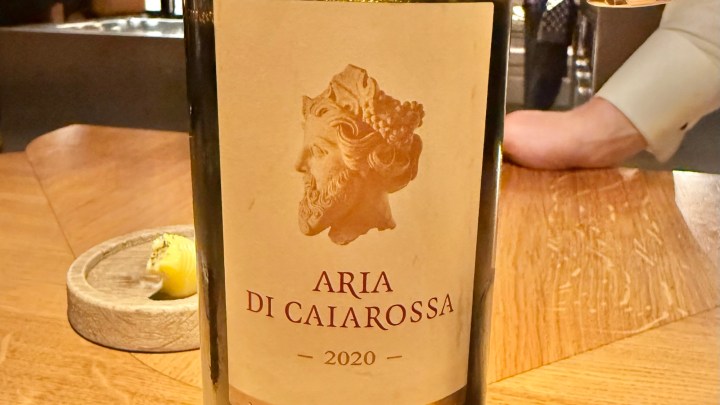 A bottle of 2020 Aria di Caiarossa wine, featuring a detailed label with a classical sculpture design, placed on a wooden table with a small dish of lemon in the background.