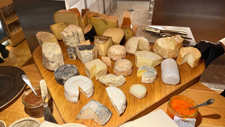 A cheese cart displaying an array of different cheeses, including various shapes and textures, on a wooden board, accompanied by a jar of spread.