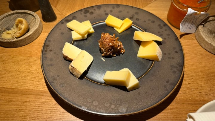 A circular plate with a variety of cheeses arranged artistically, including hard cheeses and a crispy cheese component in the center, served on a wooden table.