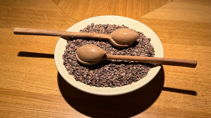 A bowl filled with cacao nibs topped with two chocolate mousse scoops served on wooden spoons.