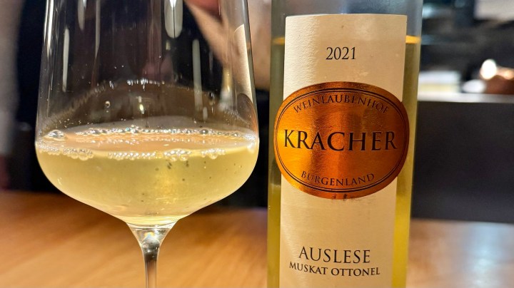 A glass of 2021 Kracher Muskat Ottonel Auslese wine alongside its bottle, placed on a wooden table.