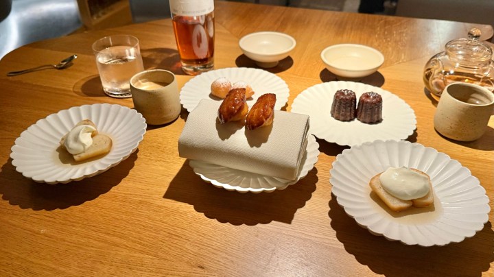 A wooden table set with various desserts on white scalloped plates, including a napkin-wrapped pastry, small cakes, and a glass of clear liquid.