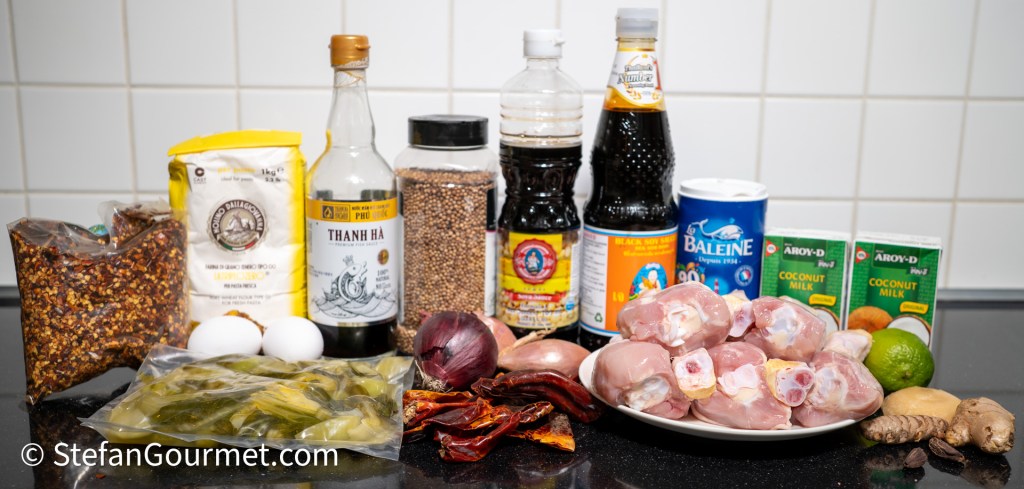 An assortment of ingredients for Khao Soi, including chicken drumsticks, coconut milk, spices, eggs, and pickled mustard greens, arranged on a countertop with a tiled background.