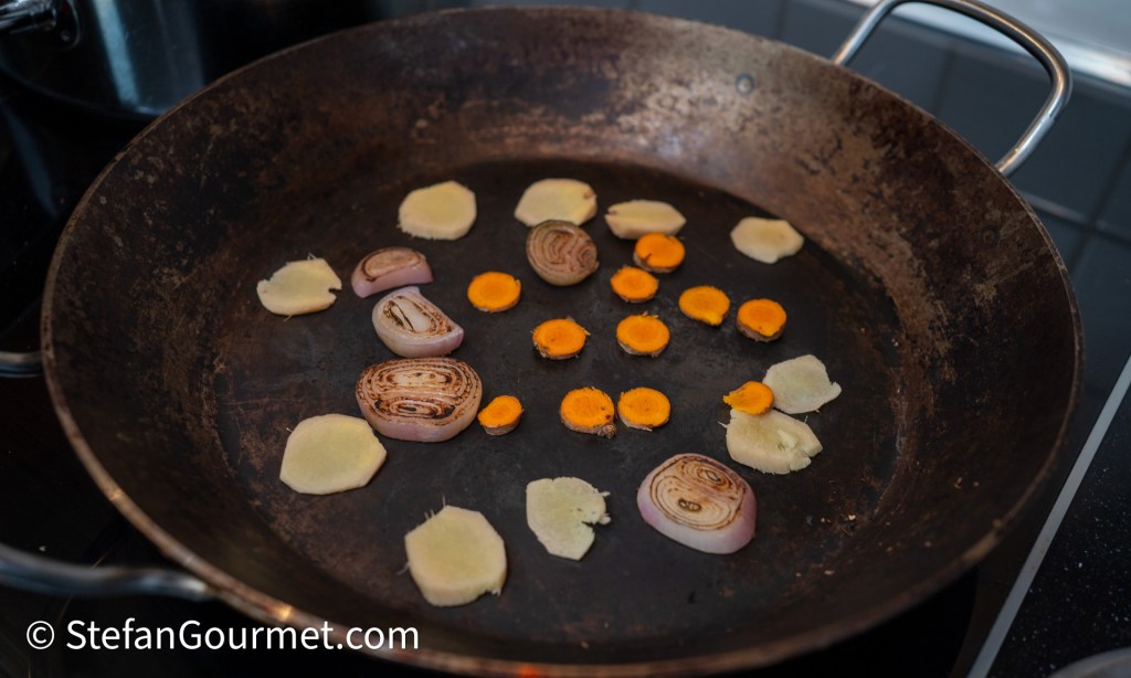 Slices of ginger, turmeric, and shallots being toasted in a metal frying pan on a stovetop.