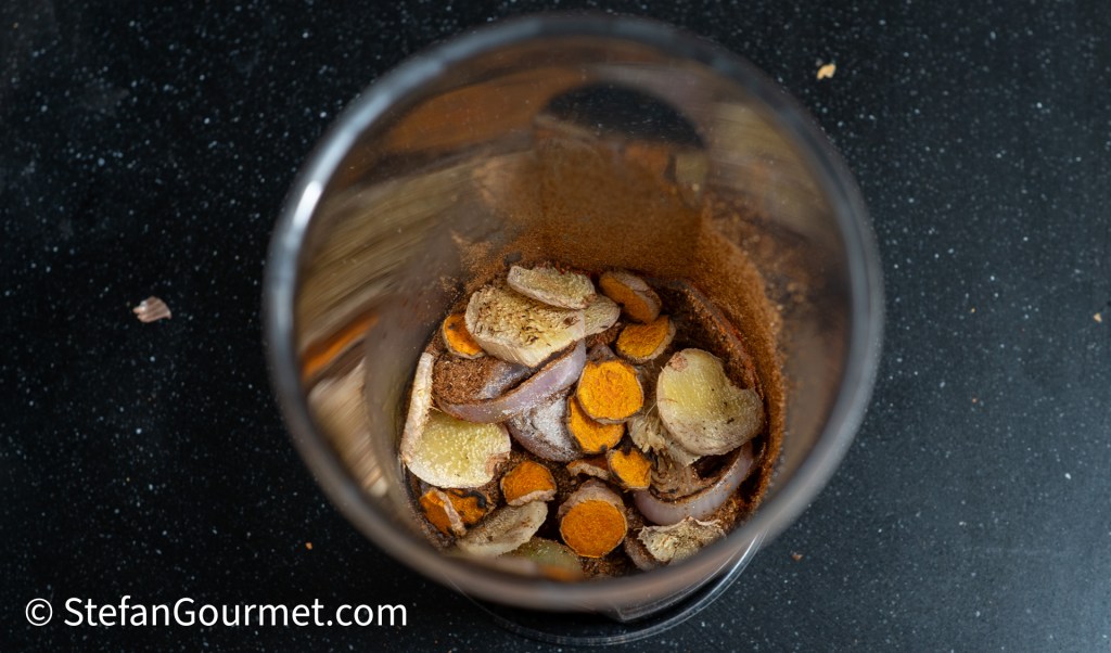 A spice grinder filled with sliced ginger, turmeric, shallots, and dried chilies, ready for grinding into a curry paste.