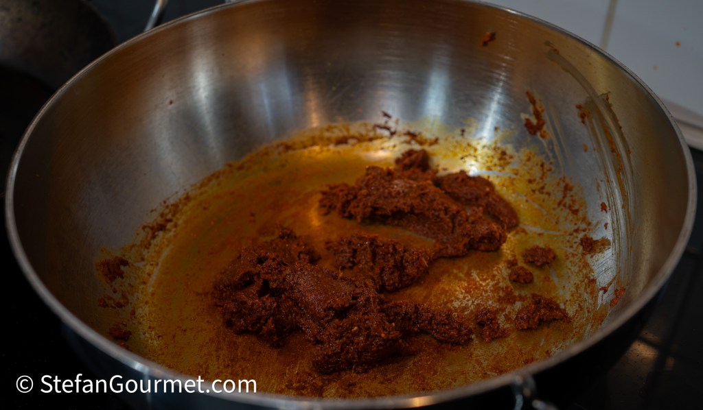 A stainless steel frying pan with a rich brown curry paste cooking at the bottom, showing remnants of oil and the vibrant color of the spices.