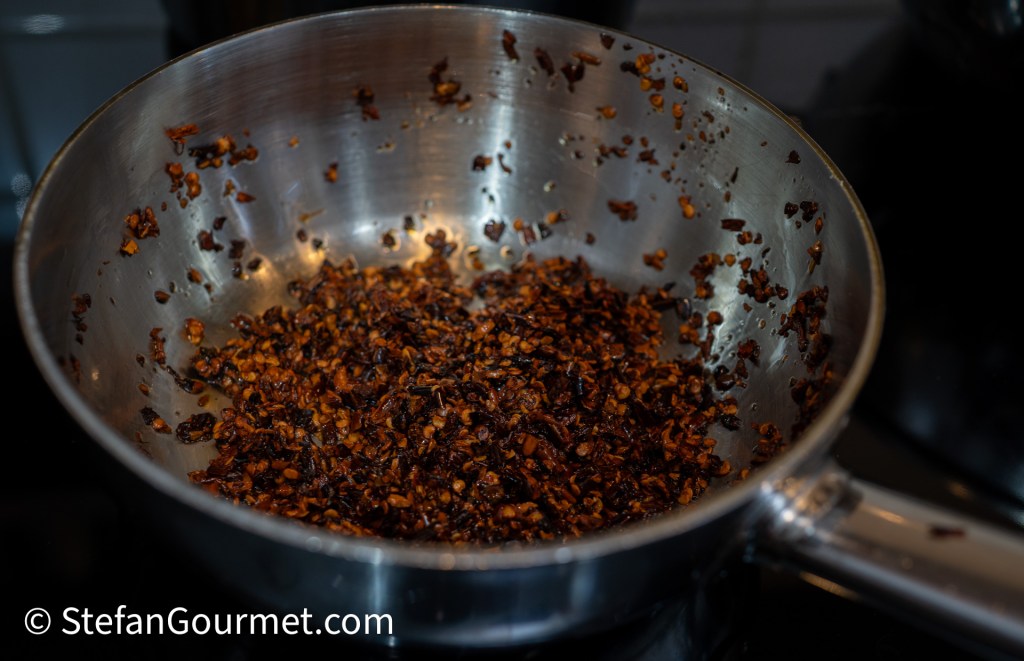 A stainless steel pan containing dark, toasted chili flakes showing a mix of various sizes, with some parts more charred, indicating they've been fried.